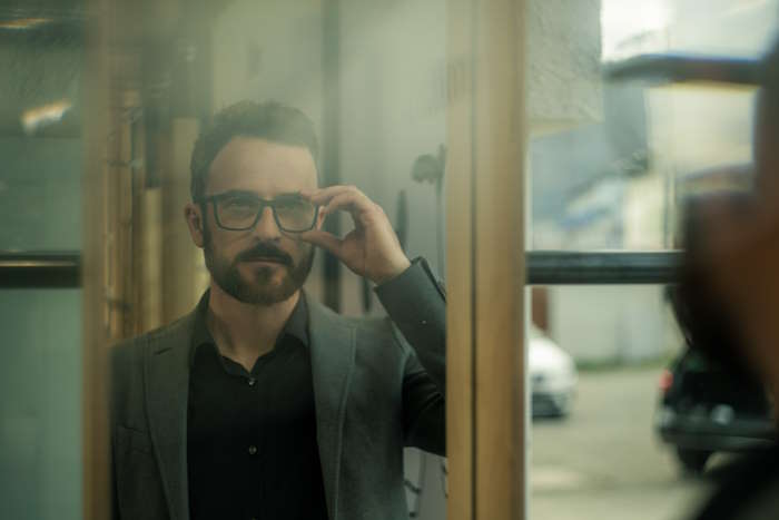 Geschäftsmann in einem grauen Anzug berührt seine Brille, symbolisch für Schmerzen hinter dem Auge, spiegelnd in einer Glasscheibe mit städtischer Unschärfe im Hintergrund.