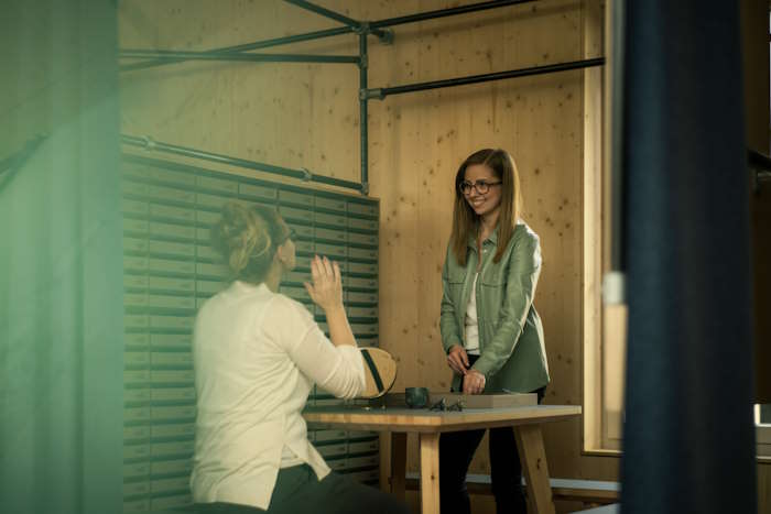 Zwei Frauen im Gespräch über Augenzucken, eine nachdenkliche Geste zeigend, in einem Büro mit Holzinterieur.