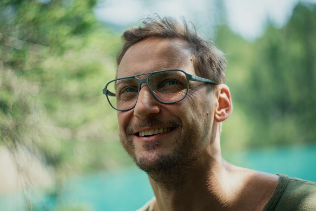 Ein fröhlicher Mann mit Brille steht vor einem grünen, verschwommenen Hintergrund, welcher das Licht sanft filtert und die Augen schont.