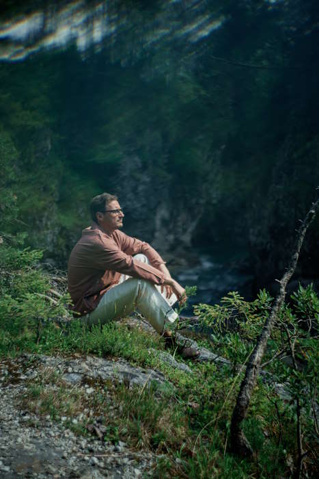 Mann in entspannter Pose sitzt inmitten der Natur mit Blick auf eine Schlucht, umgeben von Bäumen, die einen natürlichen Schutz für die Augen darstellen.