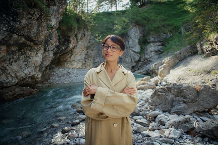 Junge Frau in einem eleganten beigen Mantel kreuzt ihre Arme und blickt durch ihre Brille auf eine natürliche Flusslandschaft, die zur Augenerholung einlädt.