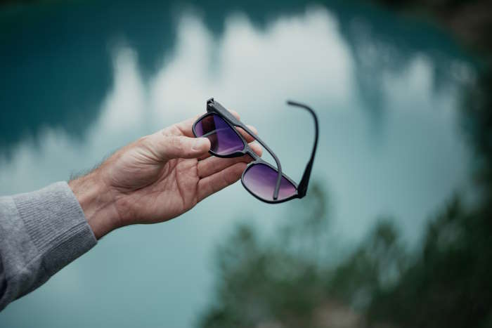 Hand hält Sonnenbrille mit reflektierenden Gläsern vor verschwommenem Bergseepanorama