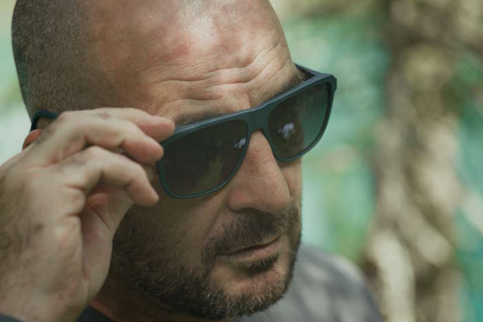 Mann mit runder Gesichtsform hebt seine dunkel getönte Sonnenbrille in einem grünen Wald an