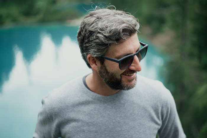 Lächelnder Mann mit trendiger Sonnenbrille mit Tönung vor einem Bergsee