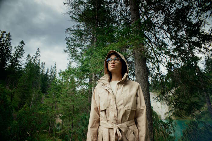 Frau in beigem Trenchcoat steht vor einem Nadelwald