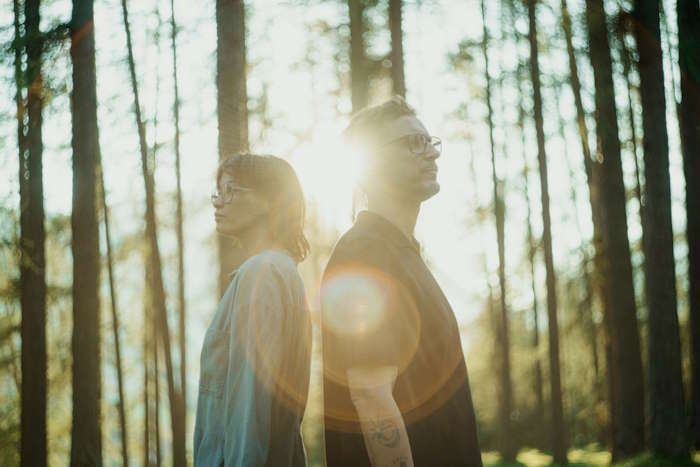 Zwei Personen mit Hornbrillen stehen Rücken an Rücken im Wald bei Sonnenuntergang