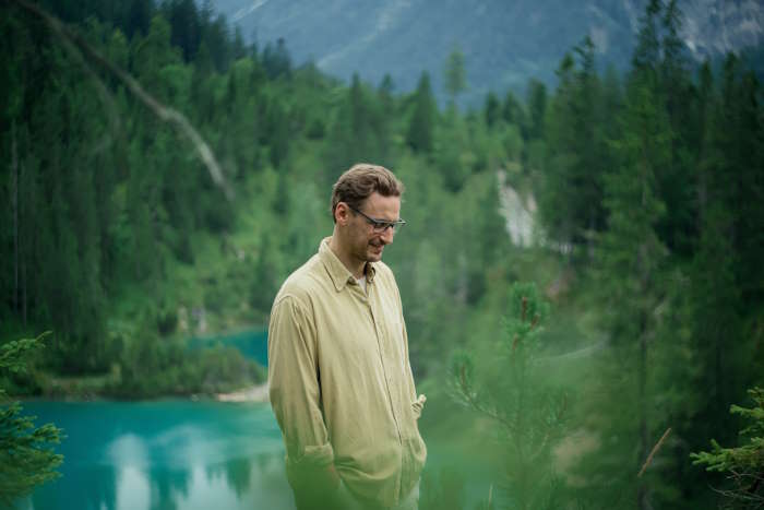 Ein Mann im beigen Hemd steht in einem dichten Wald mit Blick auf einen Bergsee, gelbe Augen durch Alkohol.