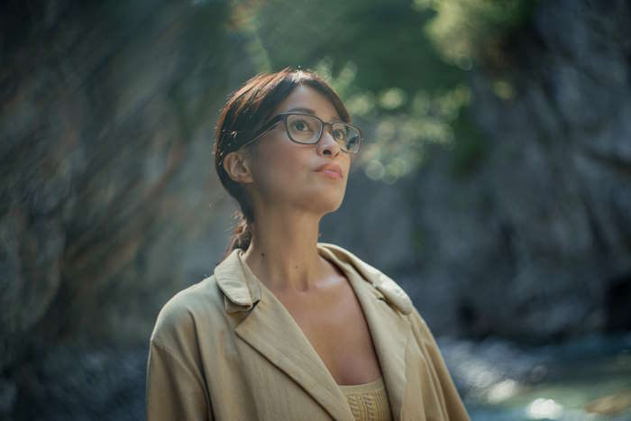 Eine Frau mit Brille steht vor einem Felsen und schaut nachdenklich in die Ferne.