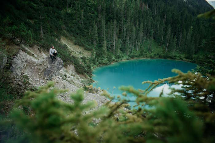 Ein Mann sitzt auf einem Felsen mit Blick auf einen türkisblauen See, umgeben von dichten Wäldern in den Bergen.