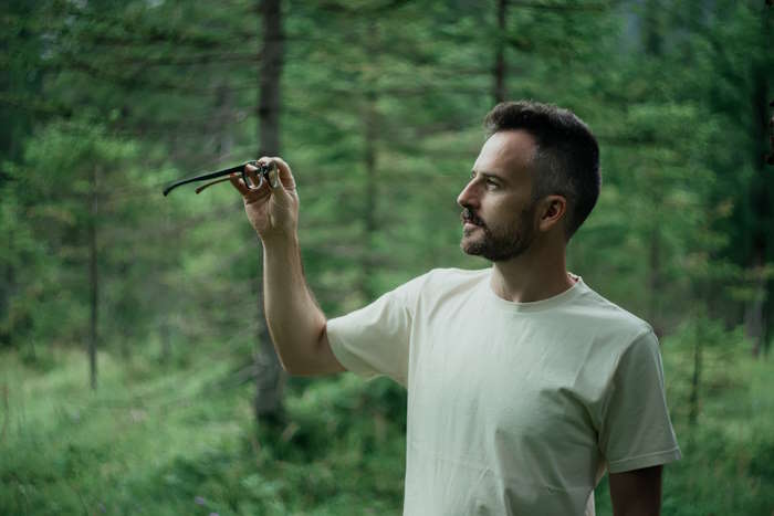 Ein Mann hält eine Brille in der Hand und schaut sie im Wald konzentriert an.