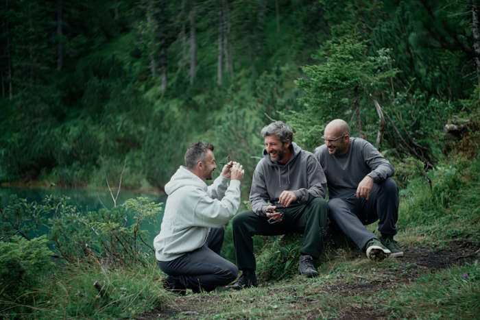Drei Männer in Outdoor-Kleidung sitzen und knien auf einer Wiese im Wald und lachen miteinander