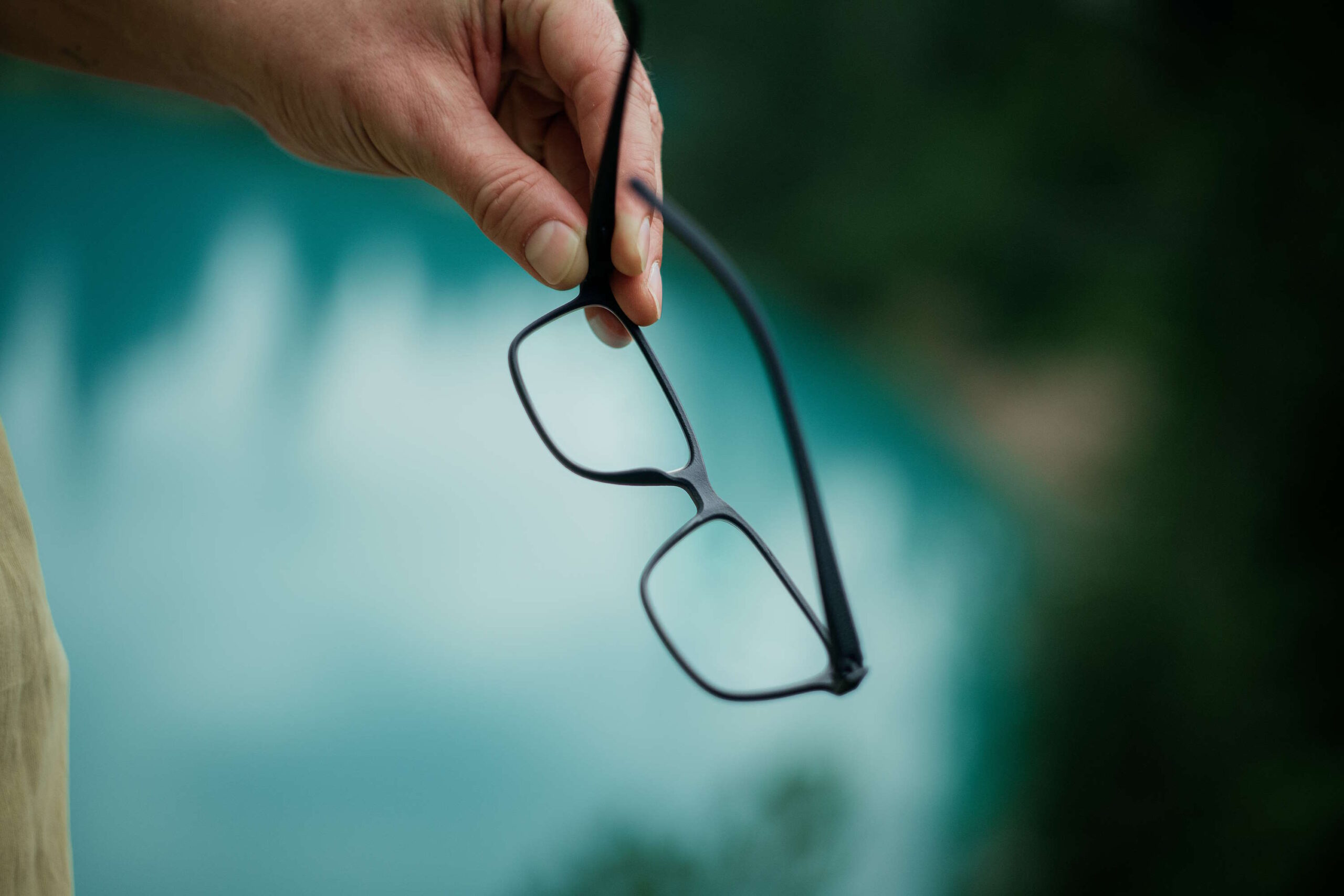 Nahaufnahme einer Hand, die eine schwarze Brille hält, als Lesehilfe über einem verschwommenen, blauen Hintergrund.