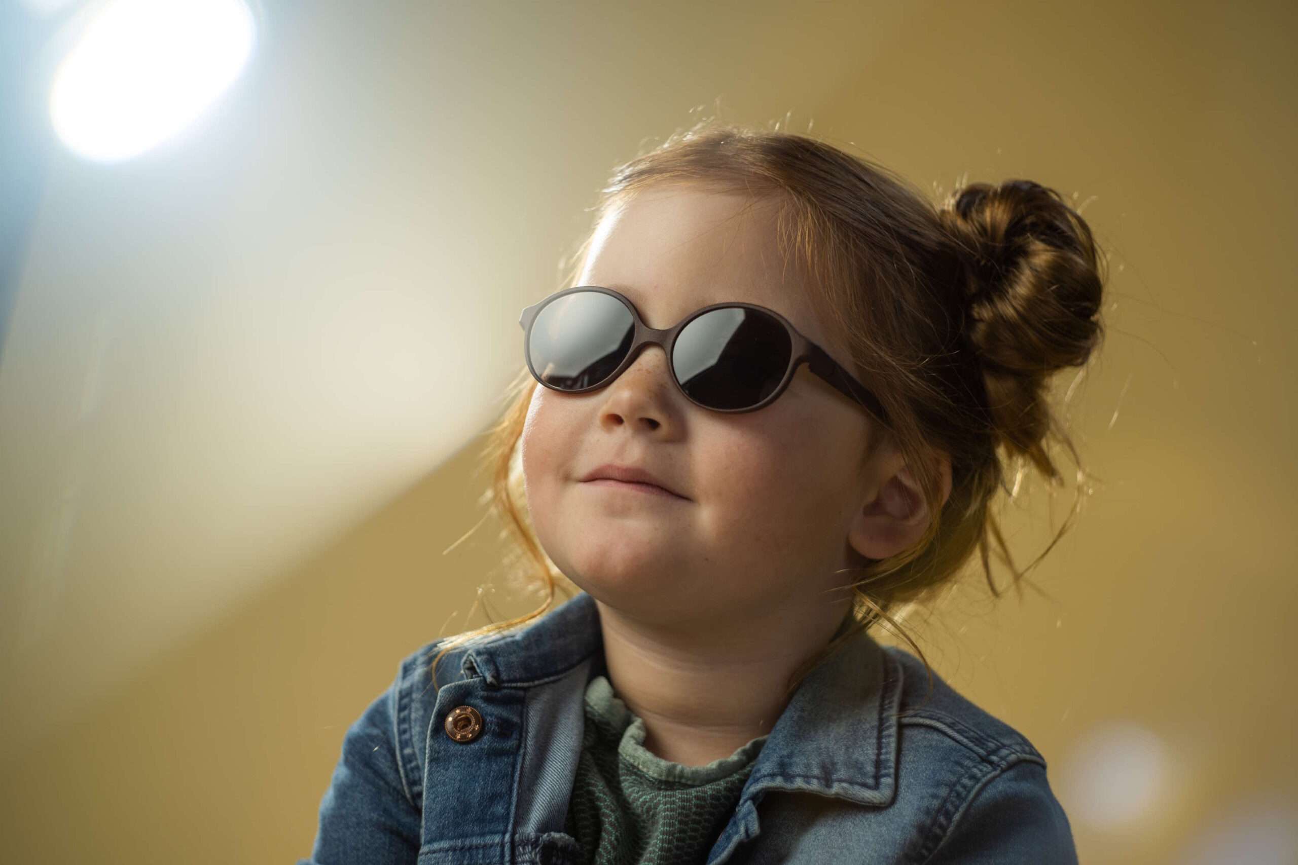 Ein kleines Mädchen trägt eine Sonnenbrille für Kids und eine Jeansjacke, während es glücklich in die Ferne schaut.