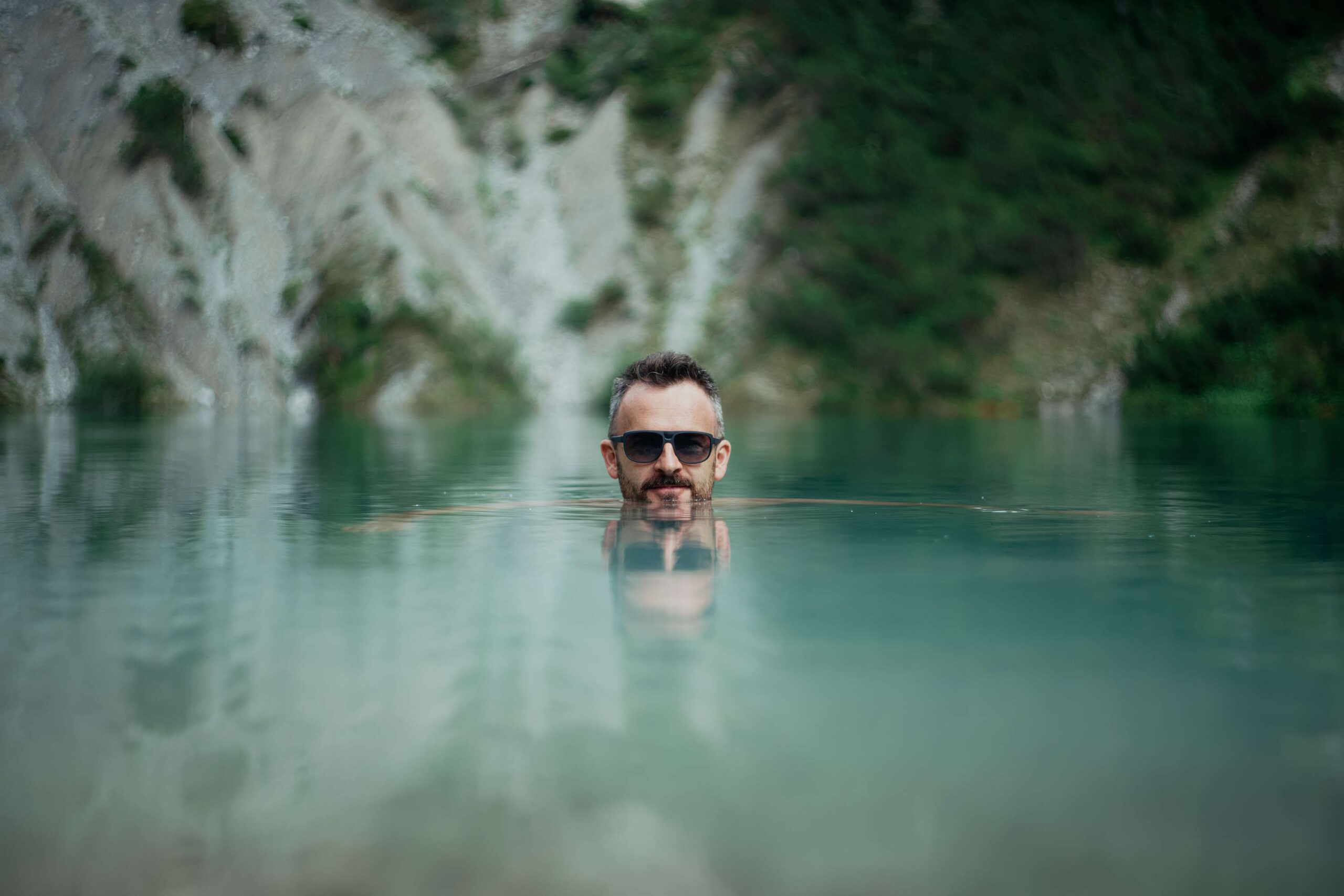 Mann mit Sonnenbrille schwimmt im klaren Wasser eines Sees, felsige Landschaft im Hintergrund - UV bedeutet
