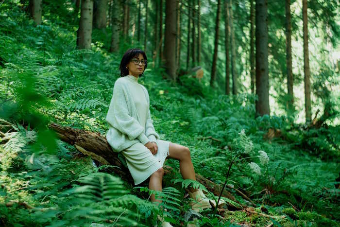 Frau mit modischer Damenbrille für den Sommer in hellem Outfit sitzt im Wald auf einem Baumstamm.