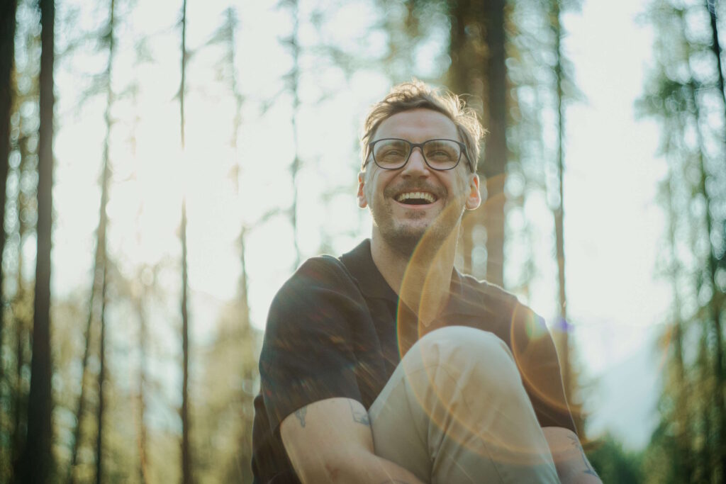 Mann mit Brille lacht im Wald bei Sonnenschein mit einer seiner Männerbrillen für den Sommer.