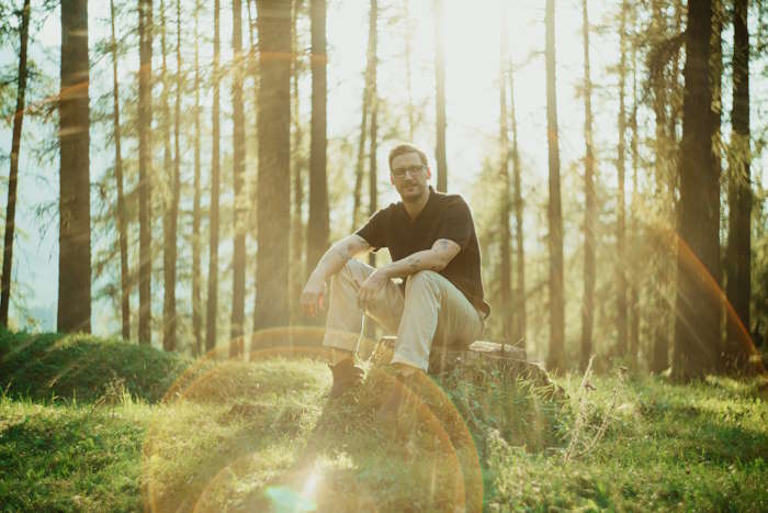 Ein Mann sitzt im Sonnenlicht auf einem Baumstumpf, die Männerbrille für den Sommer lässig getragen.