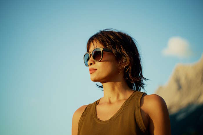 Porträt einer jungen Frau mit Sonnenbrille im Freien, die das warme Sonnenlicht genießt, während sie ihre Augen und die empfindliche Haut darunter vor Augenringen schützt.