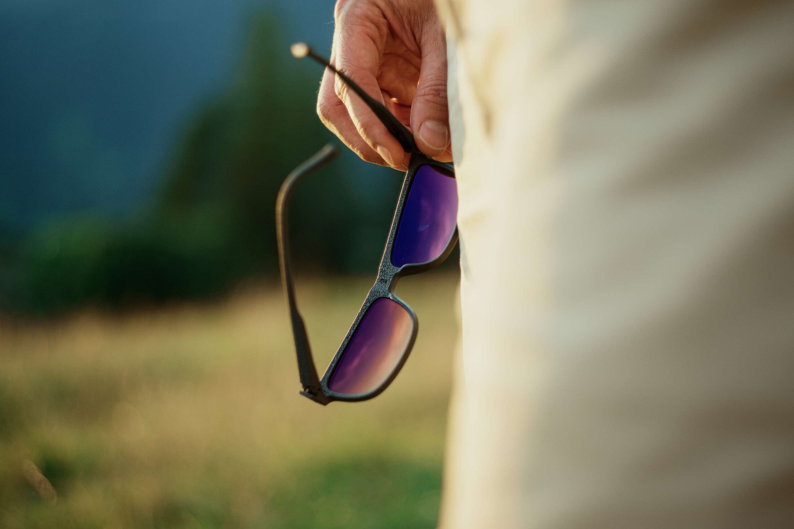 Nahaufnahme einer Hand, die eine Brille mit violett getönten Gläsern im Freien hält, symbolisch für den Schutz vor Augenringen durch Sonnenlicht.