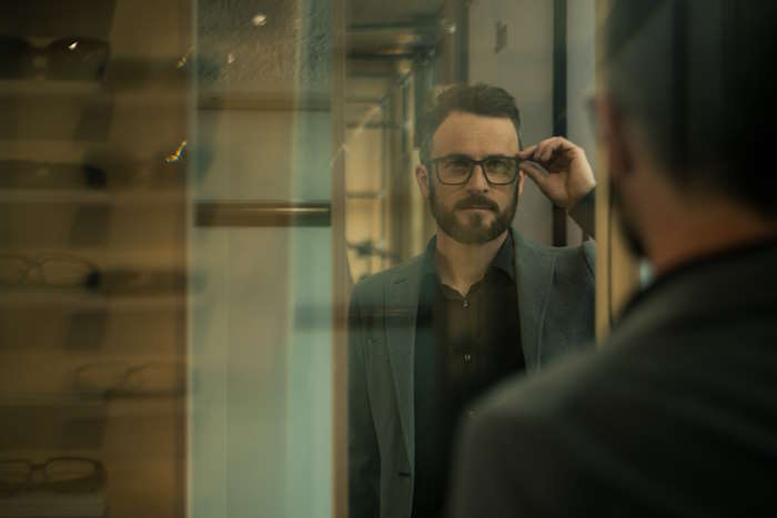 Ein Mann in einem Anzug betrachtet sich im Spiegel, während er eine Brille anprobiert. Diese Brille ist ideal für Abenteurer.