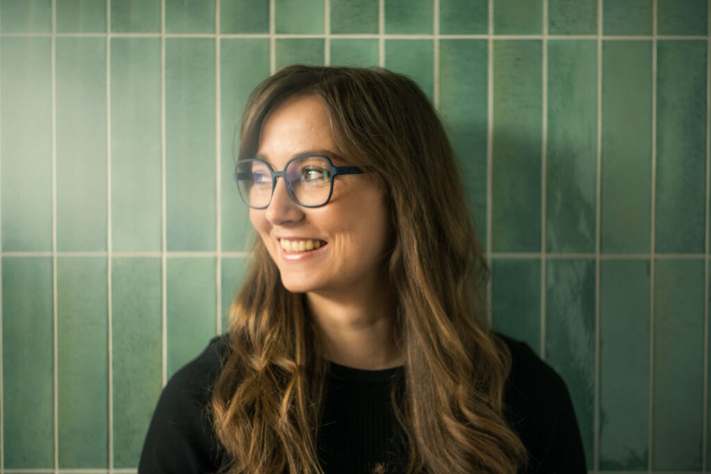 Lächelnde Frau mit modischer Brille vor einer grünen Kachelwand, perfekt für Abenteurer.