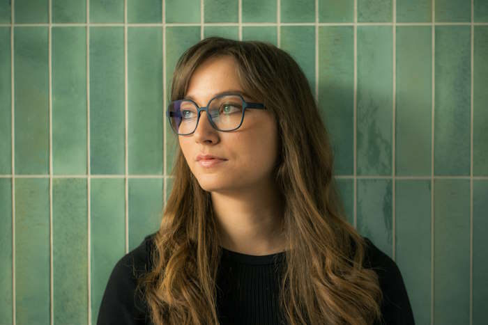 Frau mit langen braunen Haaren trägt eine modische Brille vor einer grünen Kachelwand, passend für Abenteurer.