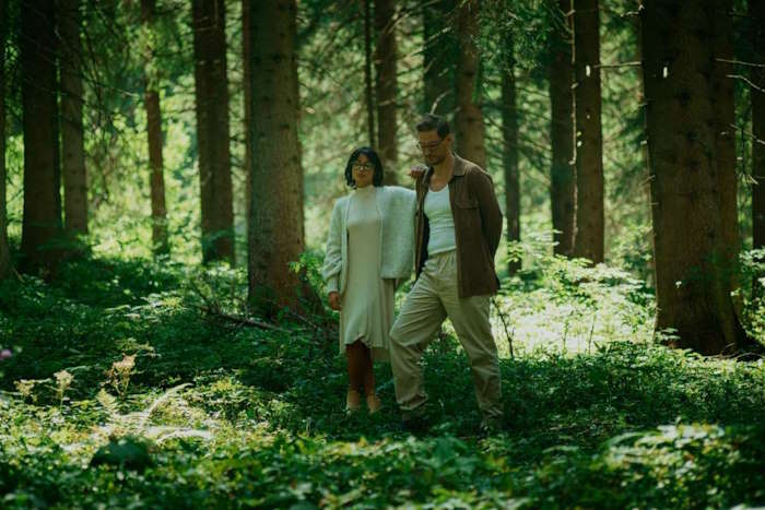 Ein Paar, bestehend aus einer Frau mit Brille und Augenringen sowie einem Mann, spaziert durch einen dichten, grünen Wald, umgeben von hohen Bäumen.