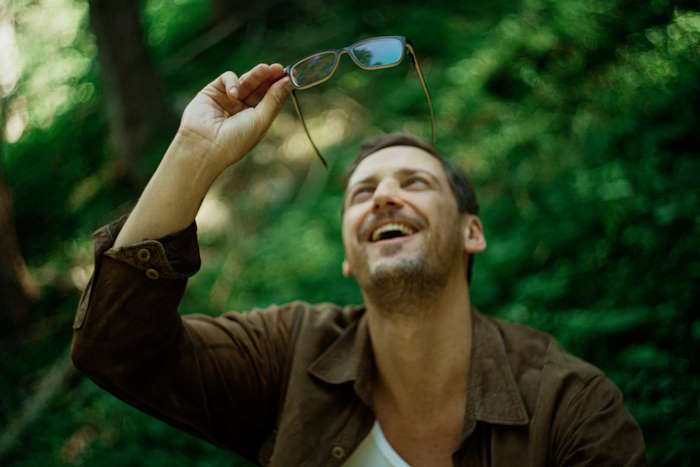 Lachender Mann im Freien, der seine Brille hochhält und in den Himmel blickt, umgeben von grünem Wald; leichte Augenringe deuten auf Müdigkeit hin.