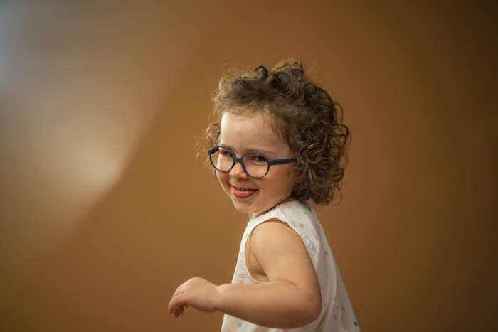 Kleines Kind mit Brille im Kindergarten, lächelnd vor braunem Hintergrund.