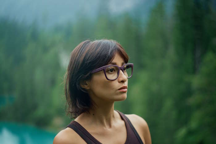 Frau mit lilafarbener Brille steht in der Natur und überlegt, welche Brillenfarbe zu welchem Typ passt.