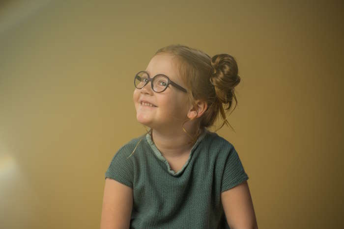 Lächelndes Mädchen mit Brille im Kindergarten vor einem neutralen Hintergrund.