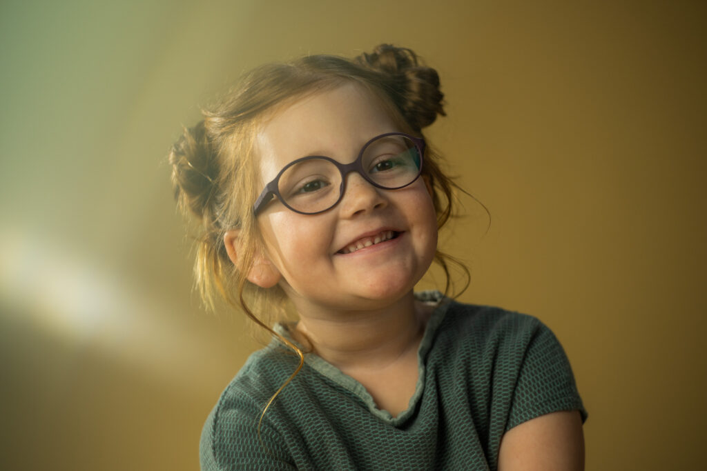 Lächelndes Mädchen mit Brille im Kindergarten vor einem neutralen Hintergrund.