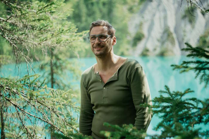 Mann mit Brille und grünem Oberteil steht lächelnd in einem Wald am türkisblauen See, umgeben von Bäumen und Natur, leicht geschwollene Augen.