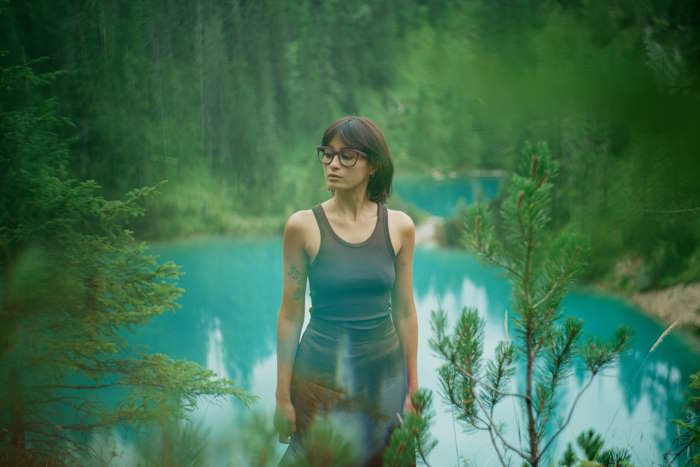 Frau in grauem Top und geschwollenen Augen steht im Wald mit Blick auf einen türkisblauen See, umgeben von Kiefern und sanften Hügeln.