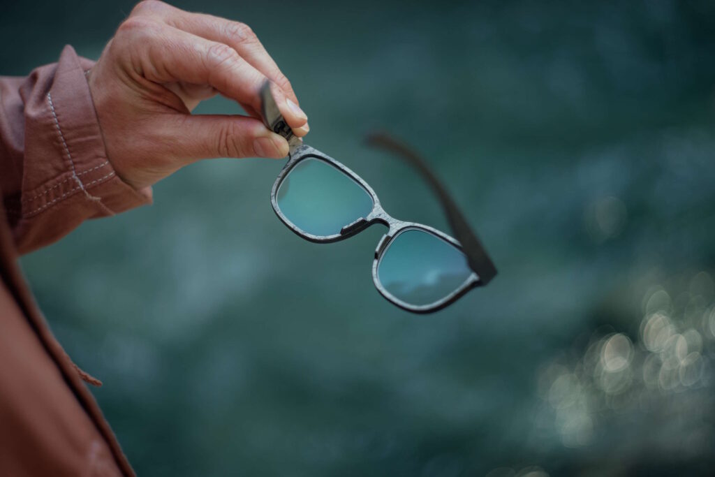 Nahaufnahme einer Hand, die eine kantige Brille für runde Gesichter hält, vor einem verschwommenen grünen Hintergrund