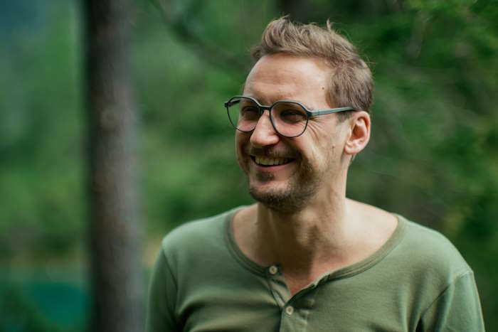 Eine lächelnde Person mit Rolf Brille in einem grünen Wald, umgeben von dichter Vegetation; die Farbe Grün prägt die gesamte Szenerie und schafft eine ruhige Atmosphäre.