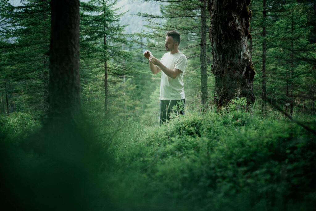 Eine Person in weißem T-Shirt fotografiert in einem grünen Wald, umgeben von Bäumen und Vegetation; die Farbe Grün gibt dem Bild eine beruhigende Note.