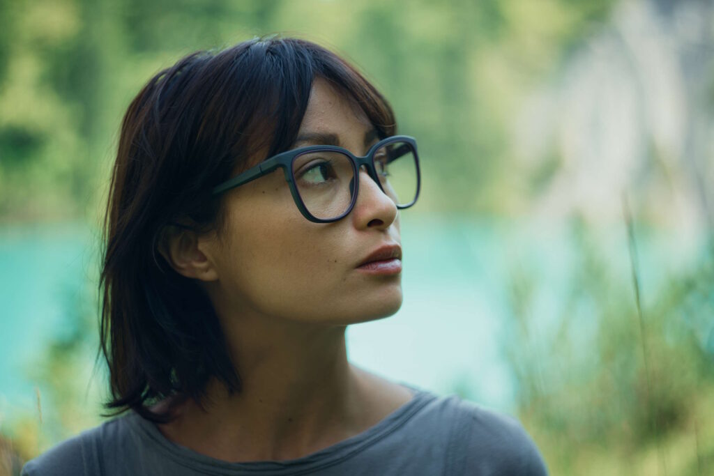 Eine Frau mit dunkler Brille schaut nachdenklich in die Ferne, umgeben von einem grünen Wald und türkisfarbenem Wasser.