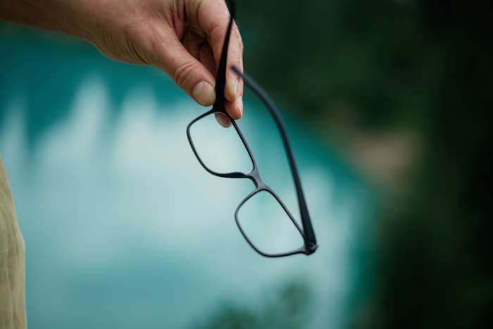 Hand hält eine Brille mit rechteckiger Augenform vor einem natürlichen Hintergrund.