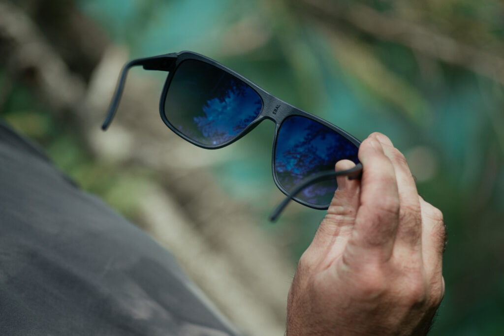 Hand hält eine Wetterbrille mit blauen Gläsern, die die Umgebung reflektieren, vor einem natürlichen Hintergrund.