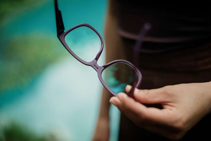 Eine Hand hält eine violette Brille vor einer verschwommenen Kulisse, ideal für den Brillen Ratgeber.