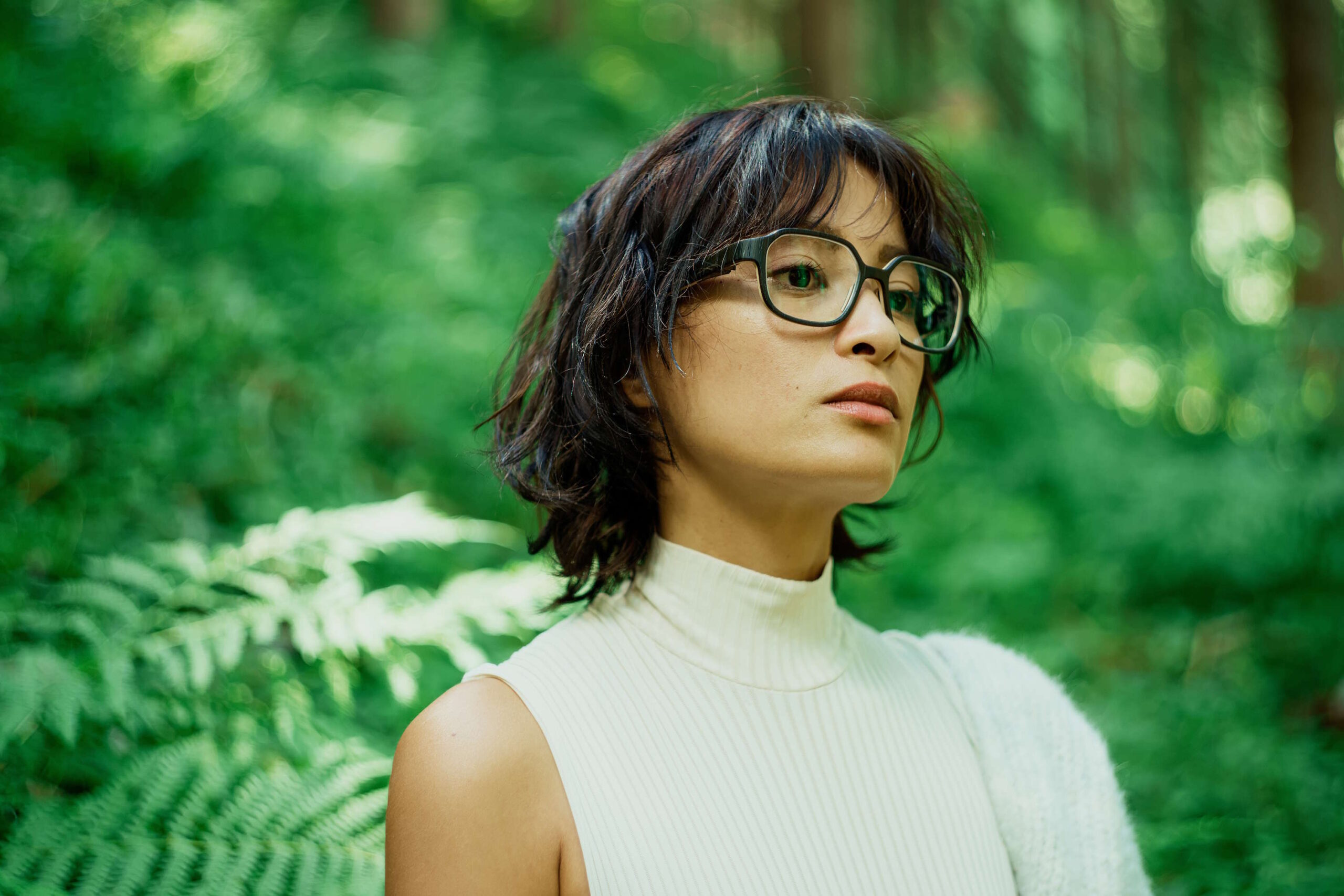 Eine Frau mit schwarzer Brille und weißem Oberteil in einem grünen Wald, ideal für den Brillen Ratgeber.