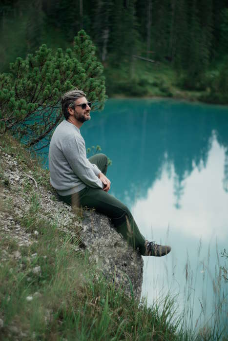 Mann mit grauen Haaren und stilvollen Brillen für graue Haare sitzt am Seeufer mit Blick ins Wasser