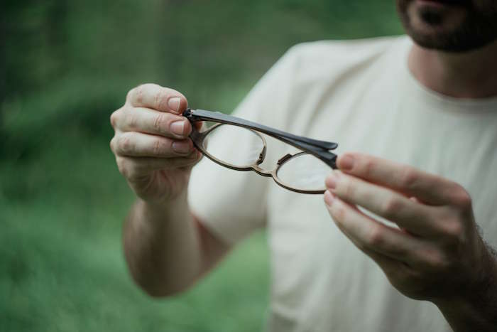 Nahaufnahme von Händen, die eine Brille im Wald halten – womöglich wegen Druck hinter dem Auge abgenommen